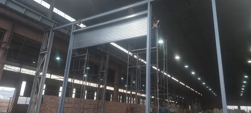 Warehouse roller door, mid-installation: A galvanised roller shutter curtain going into a newly erected steel portal frame inside a working warehouse, fitted from scaffolding at full height.