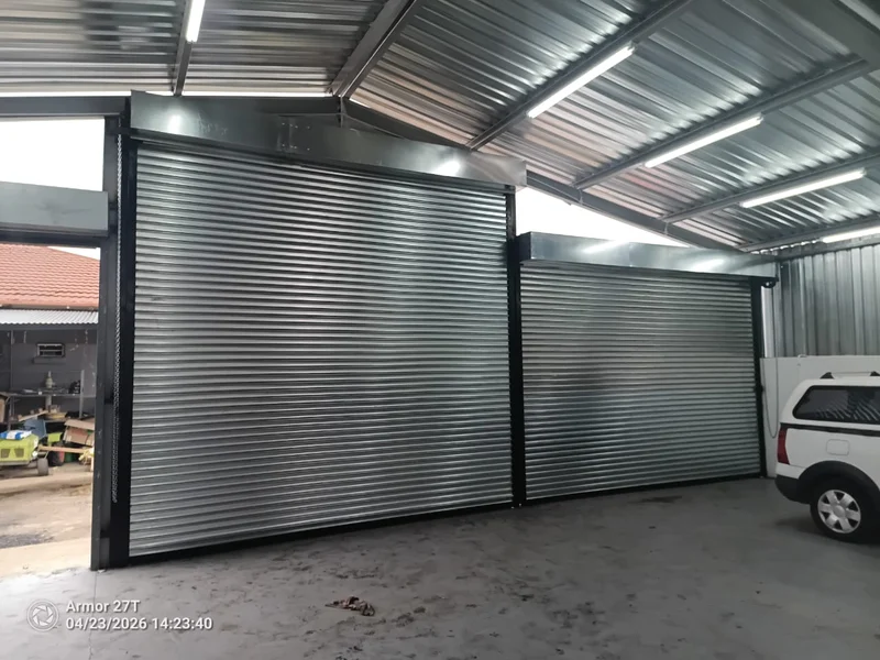 Twin roll-up doors under one roof: Two galvanised roll-up doors side by side in a steel-roofed workshop and carport, sized to two different openings.